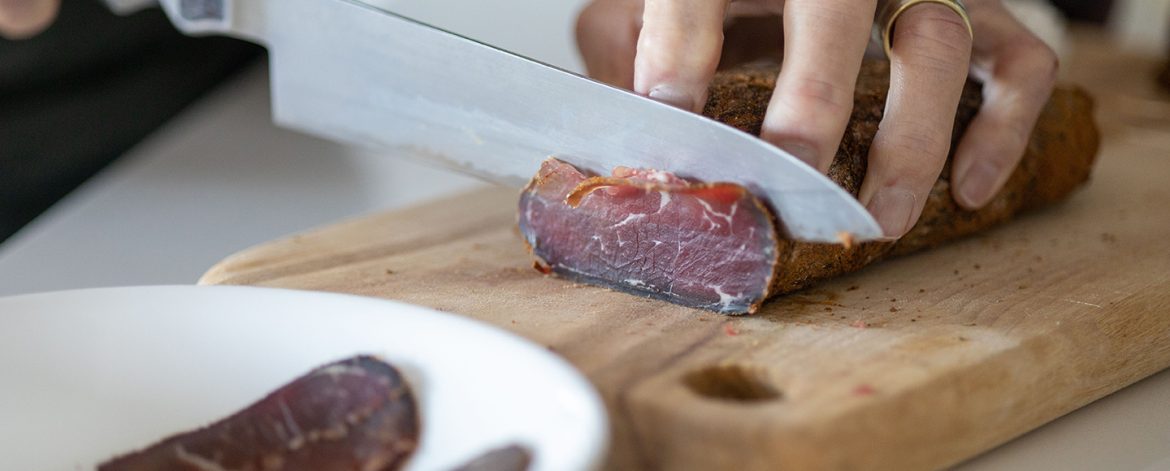 Chef preparing a meal in a kitchen, slicing pieces of pastirma on a wooden cutting board